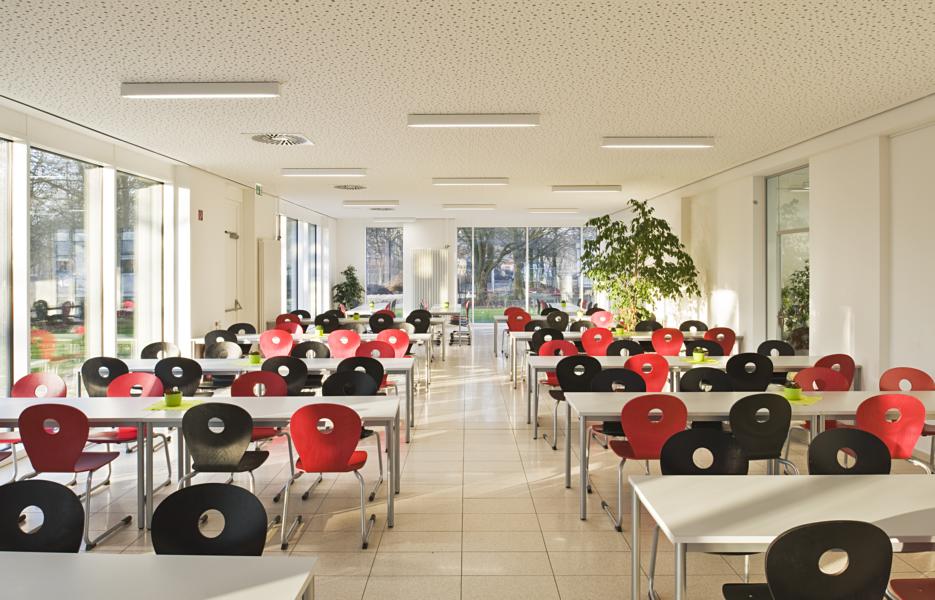 ein großer, lichtdurchfluteter Raum mit vielen roten und schwarzen Stühlen und tiefen Fenstern ist zu sehen. Es handelt sich um die Mensa der Bismarckschule.